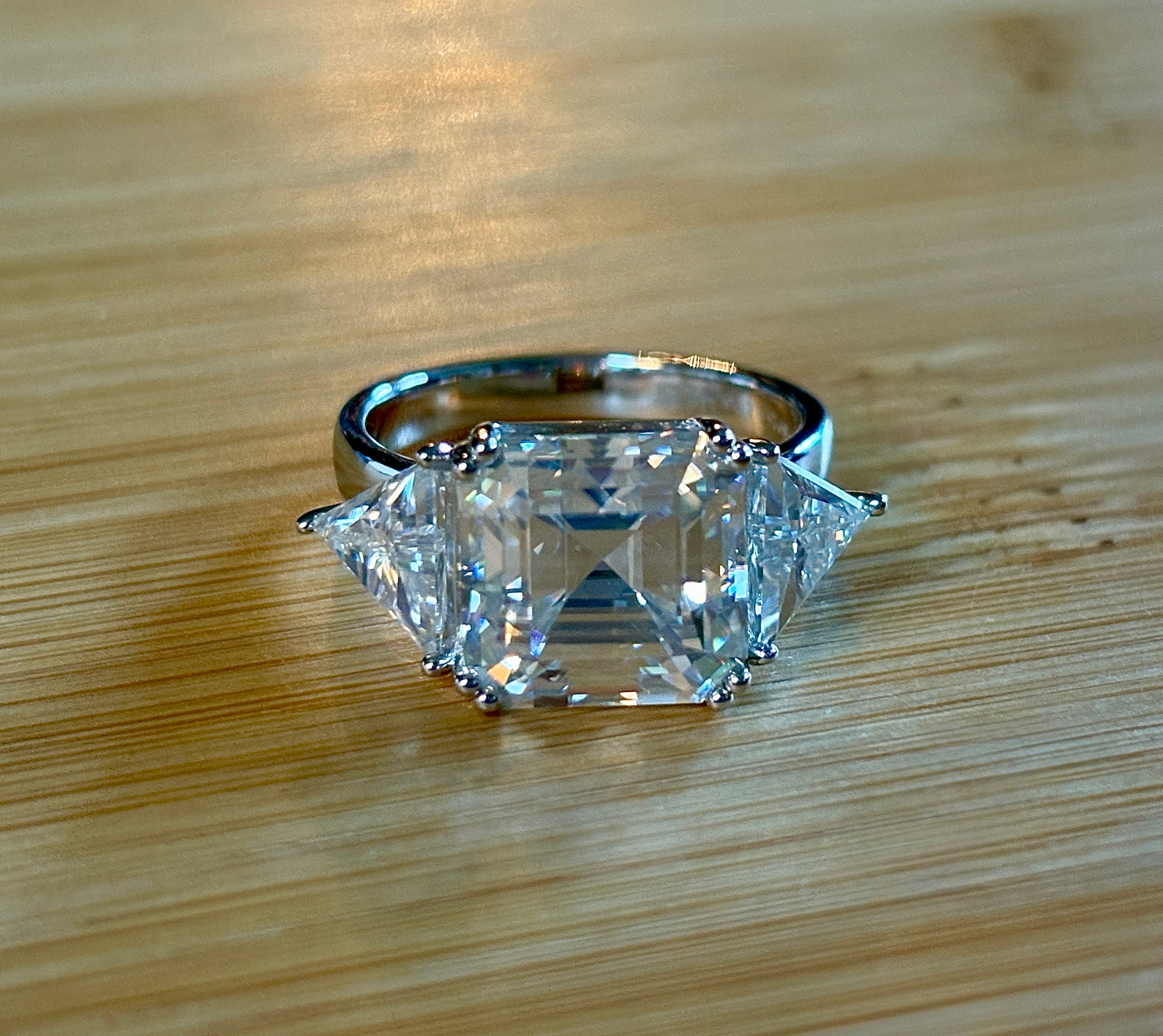 A 9.4cttw Moissanite statement ring with a 7ct Asscher-cut center stone and (2) 1.20ct Moissanite trillions, with bright 14k white rhodium!