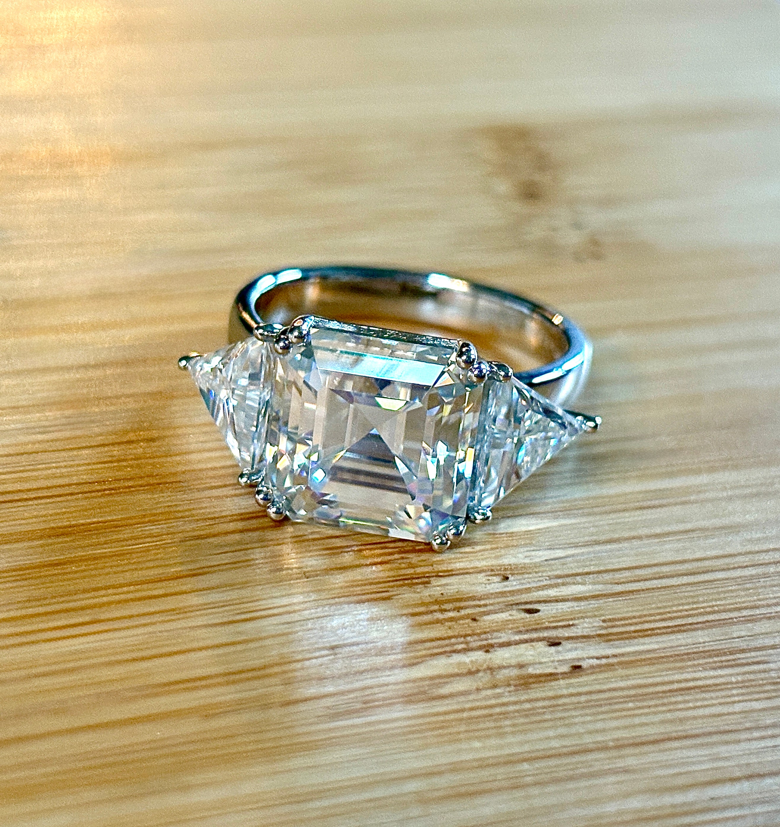 A 9.4cttw Moissanite statement ring with a 7ct Asscher-cut center stone and (2) 1.20ct Moissanite trillions, with bright 14k white rhodium!