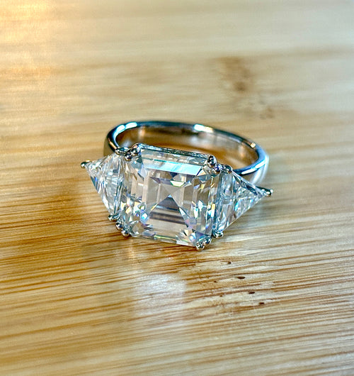 A 9.4cttw Moissanite statement ring with a 7ct Asscher-cut center stone and (2) 1.20ct Moissanite trillions, with bright 14k white rhodium!