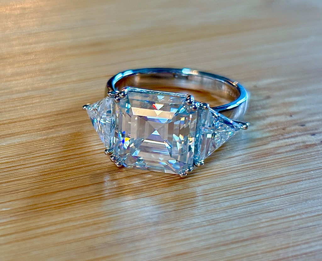 A 9.4cttw Moissanite statement ring with a 7ct Asscher-cut center stone and (2) 1.20ct Moissanite trillions, with bright 14k white rhodium!